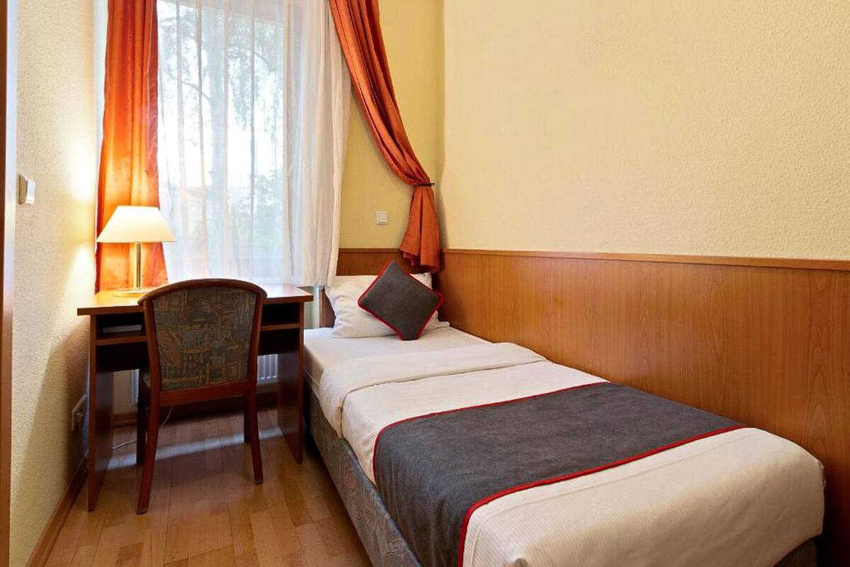 Habitación de hotel con una cama individual, un escritorio con silla, una lámpara y una ventana con cortinas.