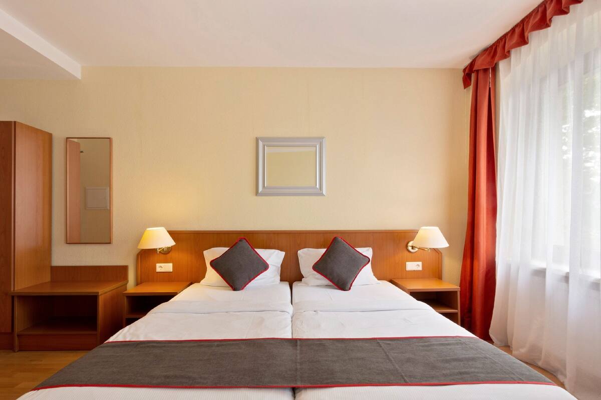 Habitación de hotel con dos camas, cabecero de madera, lámparas de noche, un espejo y una ventana con cortinas.