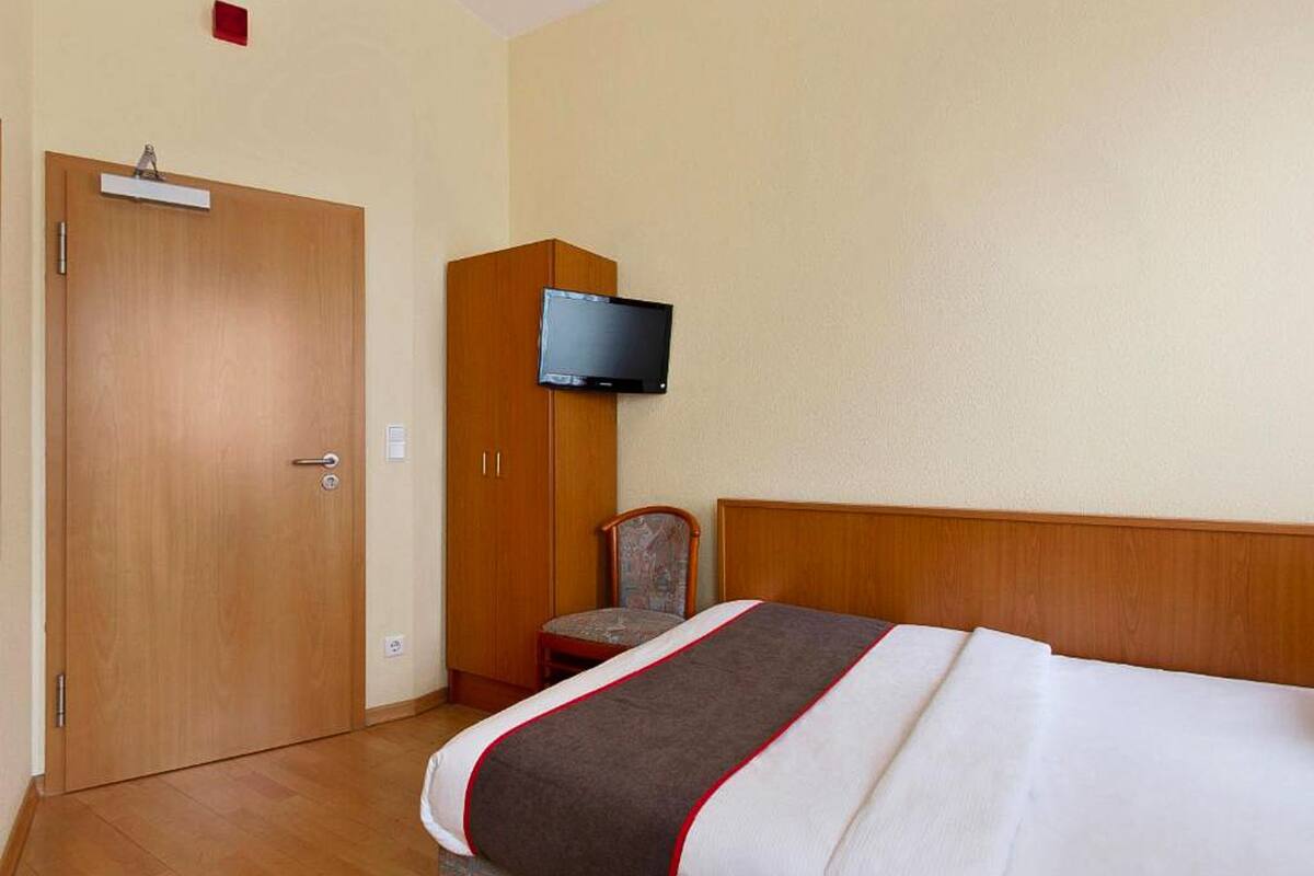 Una habitación de hotel con una puerta de madera, un televisor montado en la pared, una cama con cabecera estampada y una pequeña silla.