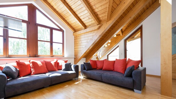 Un loft spacieux avec de hauts plafonds et des poutres apparentes, comprenant deux canapés gris avec des coussins rouges et gris, de grandes fenêtres et un plancher en bois.