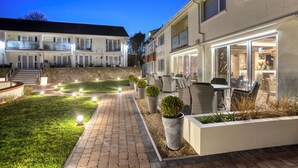 Un patio exterior bien iluminado con un sendero empedrado, plantas en macetas y mobiliario de exterior.