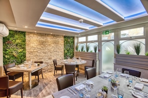 Restaurante com parede de pedra e vegetação, mesa redonda posta para refeição e teto com claraboia.