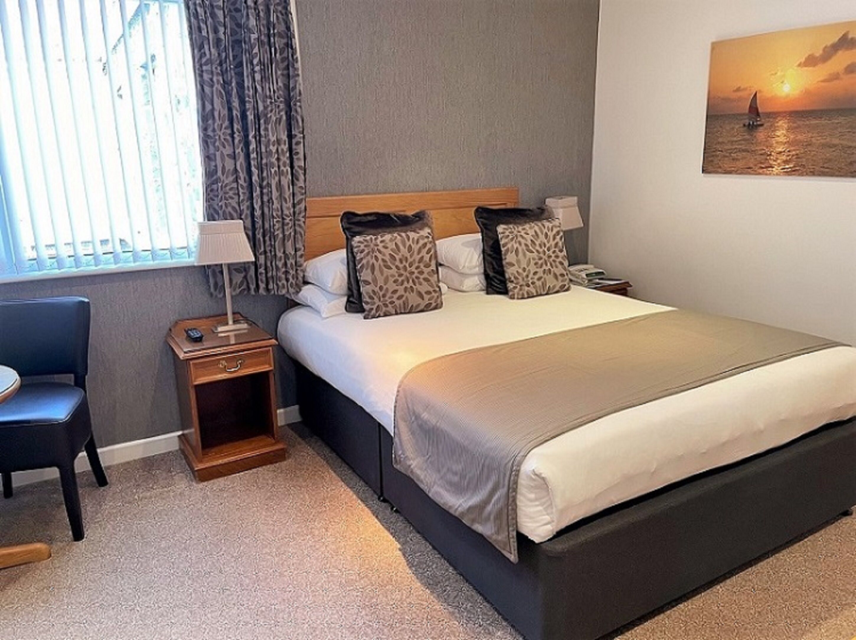 A neatly arranged hotel room with a bed, a chair, a nightstand, and a painting of a sunset over water.