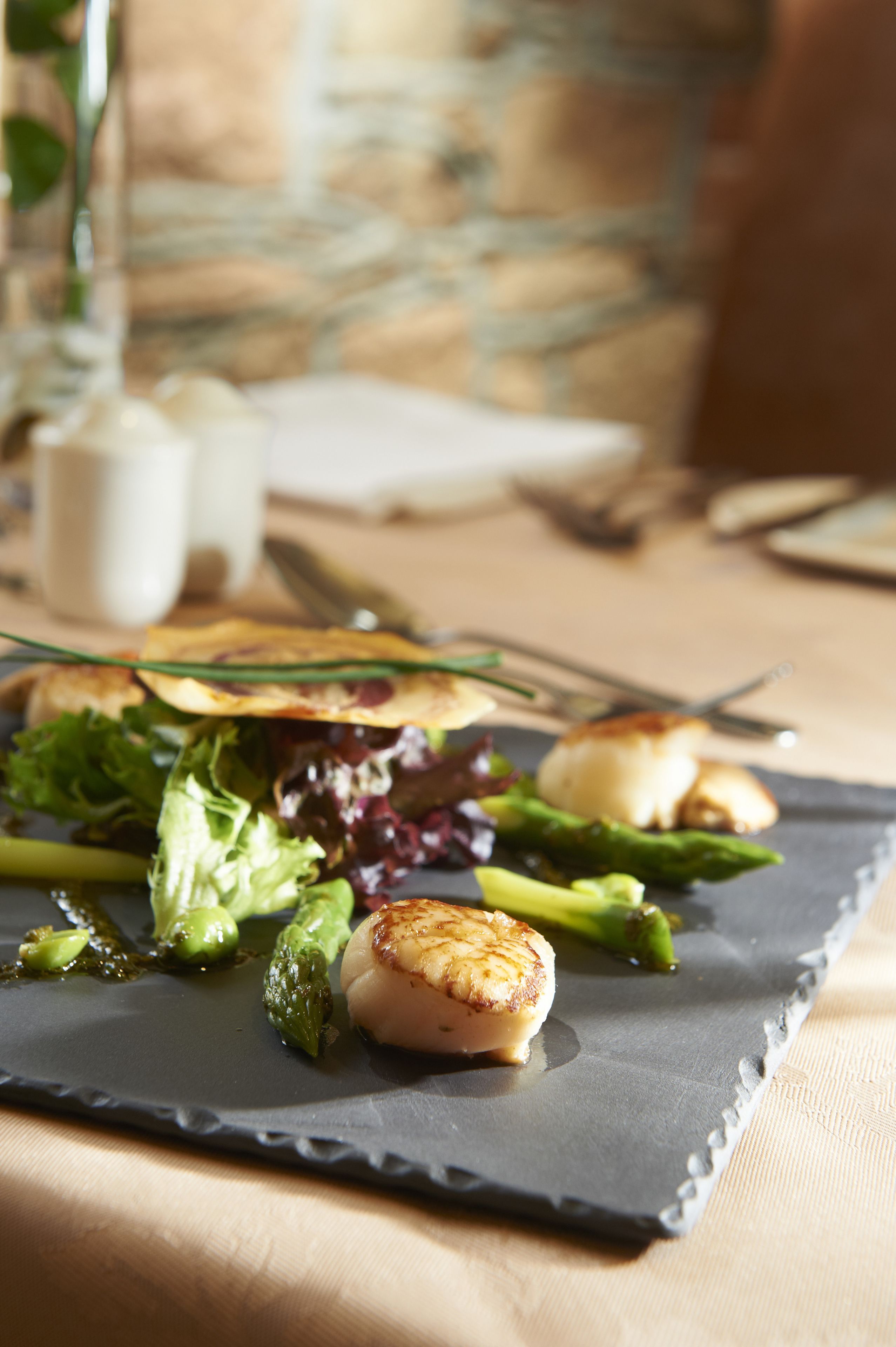 Una mesa de comedor con un plato de comida, que incluye vieiras a la plancha, espárragos y verduras de hoja, junto con una copa de vino blanco.