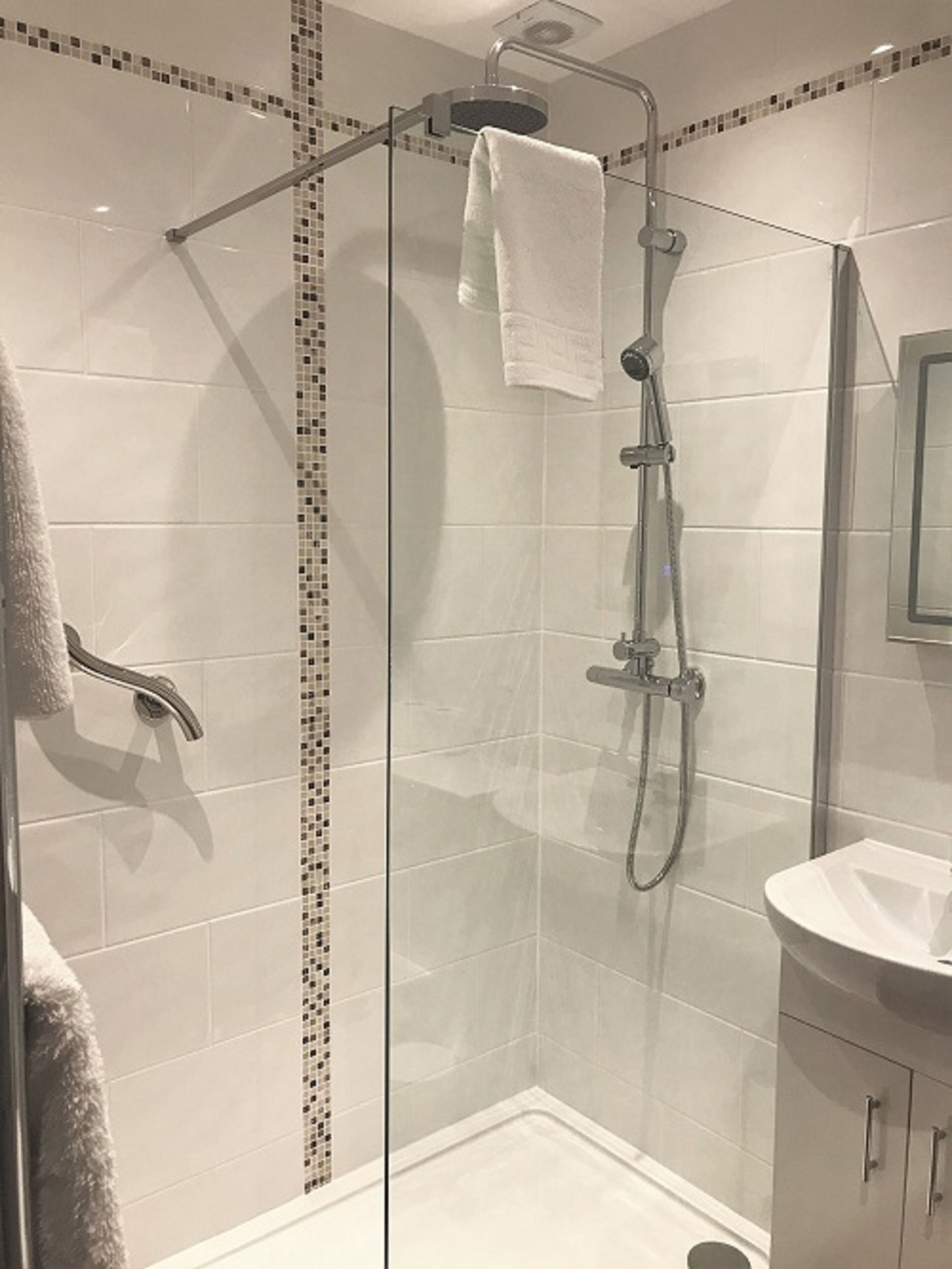 En dusch med glasväggar, en handhållen duschslang och ett fast monterat duschmunstycke ovanför, en vit handduk hängande på duschmunstycket och en vit tvättställ med en spegel ovanför.