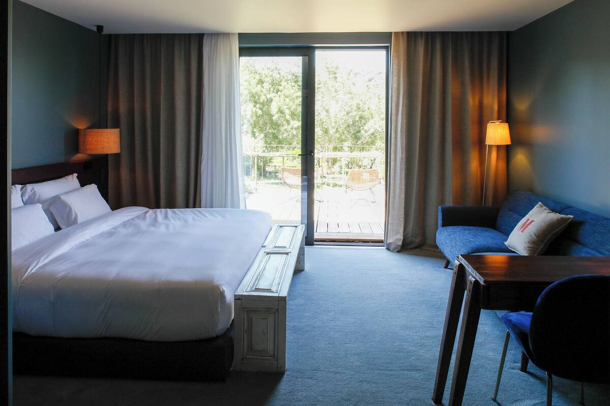 A hotel room with a large bed, a sofa, a small table, and a view of the outdoors through a sliding door.