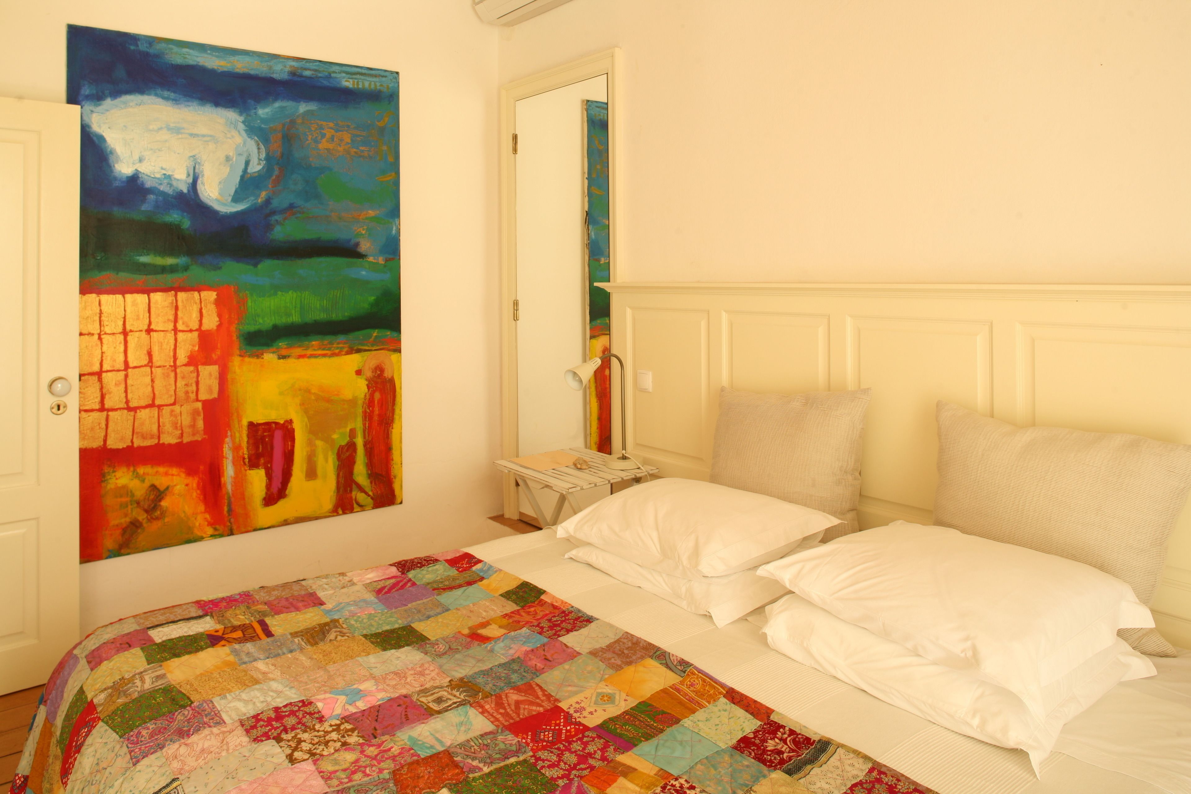 Un dormitorio con un cuadro grande y colorido en la pared, una colcha acolchada y un cabecero blanco.