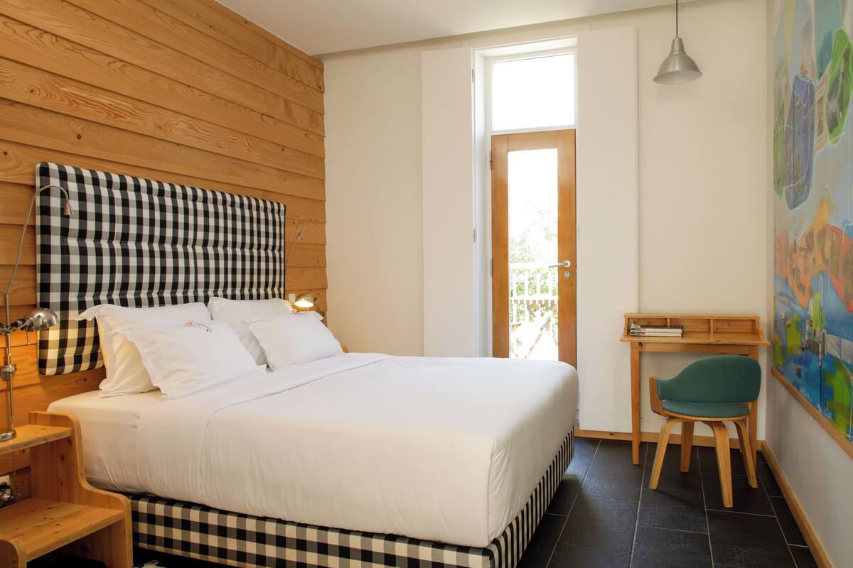 A bedroom with a large bed, a desk, a chair, and a door leading to a balcony.