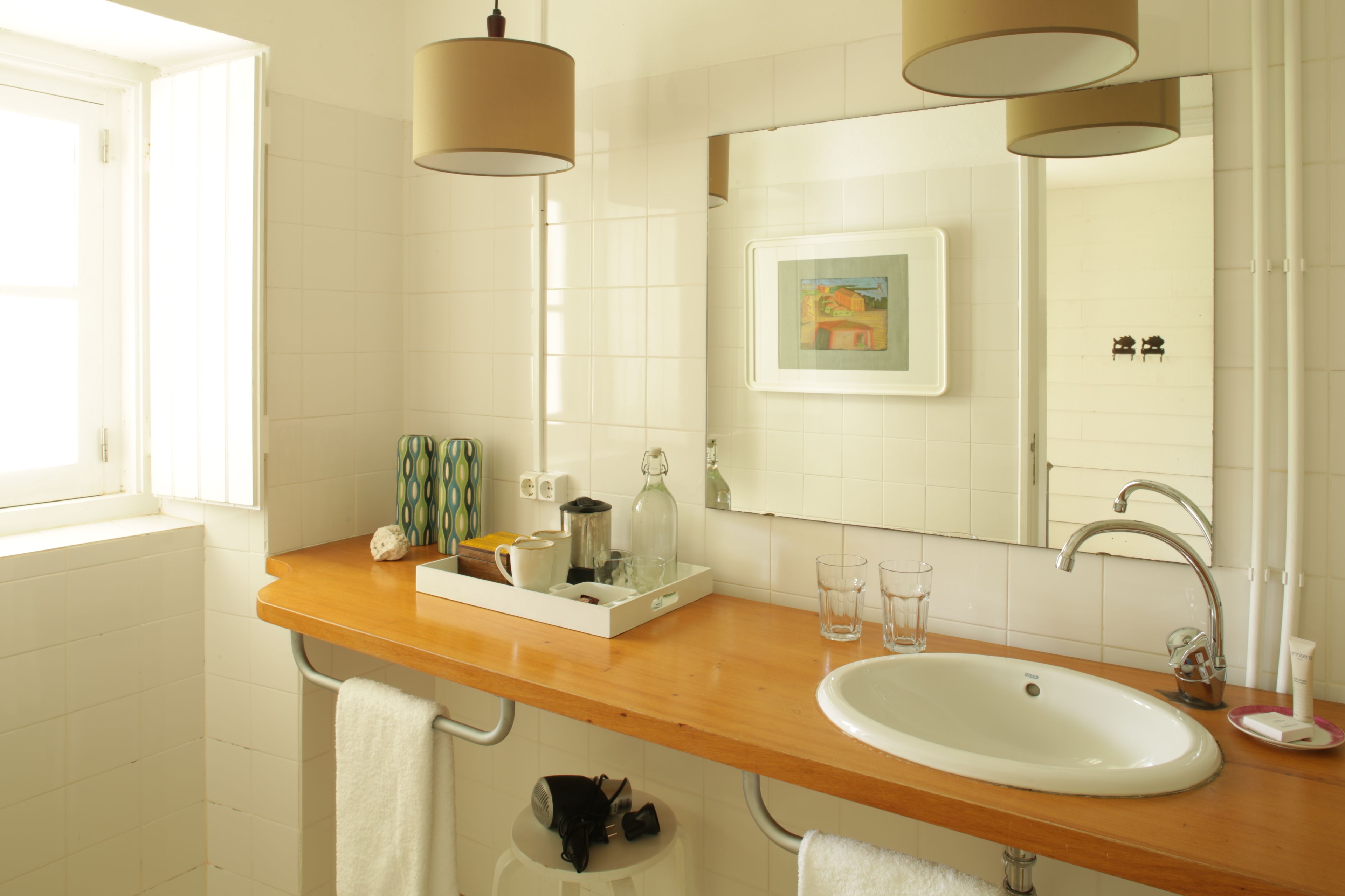 A bathroom with a wooden countertop, a white sink, and a large mirror. There are two hanging lamps, a framed picture on the wall, and a window providing natural light.