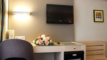 A hotel room with a flat-screen TV mounted on the wall, a desk with a glass top, a chair, a small refrigerator, and a bouquet of flowers on the desk.