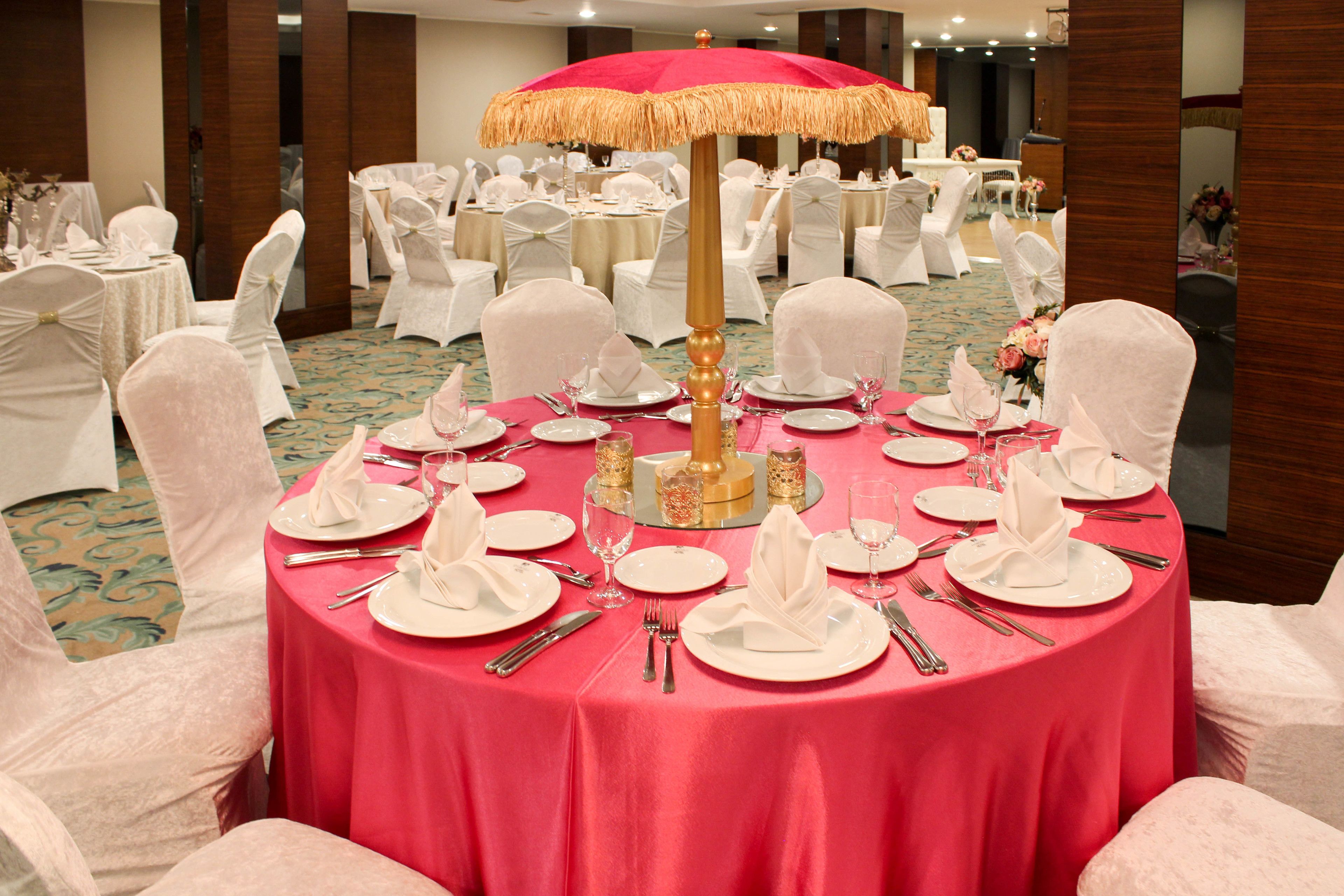 A banquet hall with round tables set for a formal event, featuring white chairs and a central table with a red tablecloth and a golden umbrella.