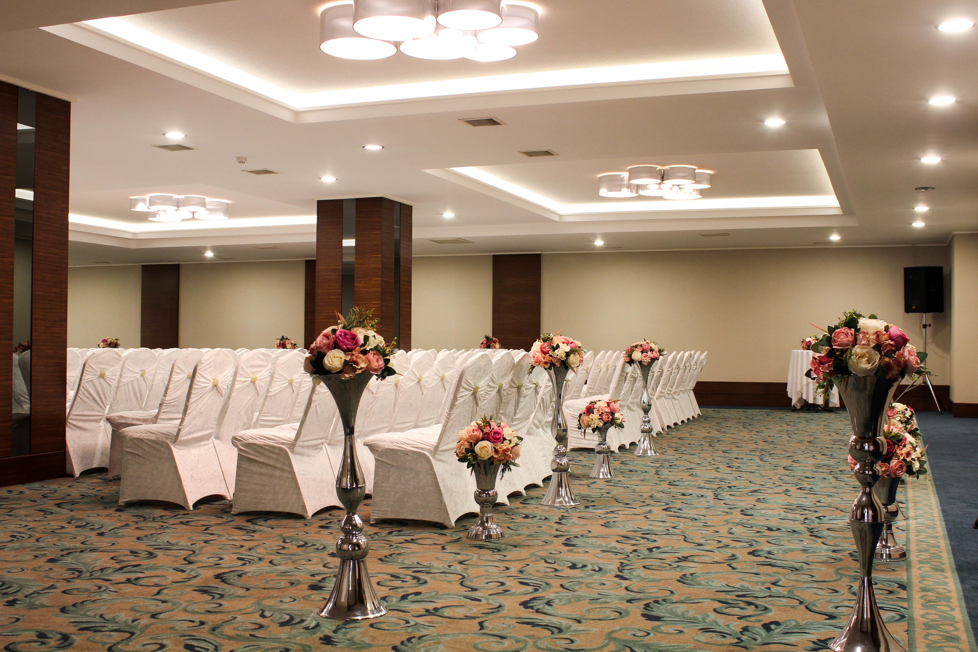 Une salle de conférence avec des rangées de chaises blanches, des arrangements floraux en guise de décoration centrale et un sol recouvert de moquette.