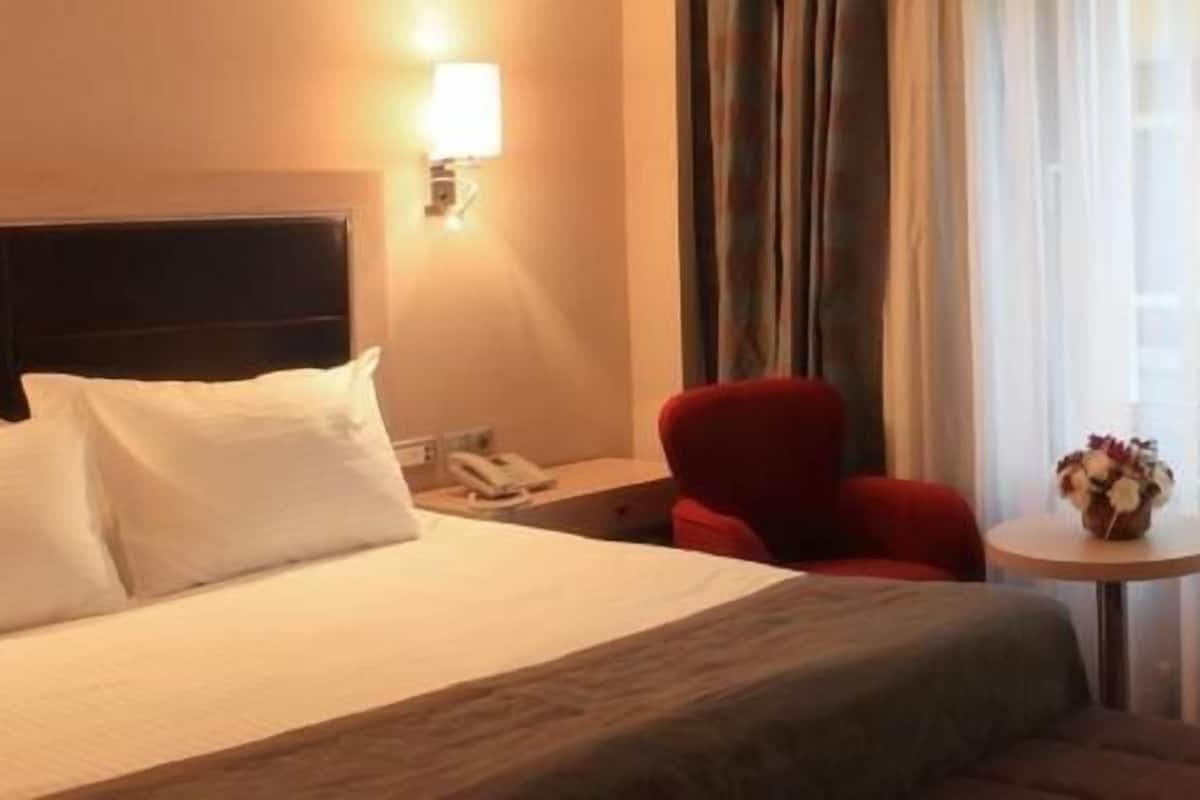 Una habitación de hotel con una cama, una mesita de noche, una silla roja, una pequeña mesa con un florero y un escritorio con un control remoto.