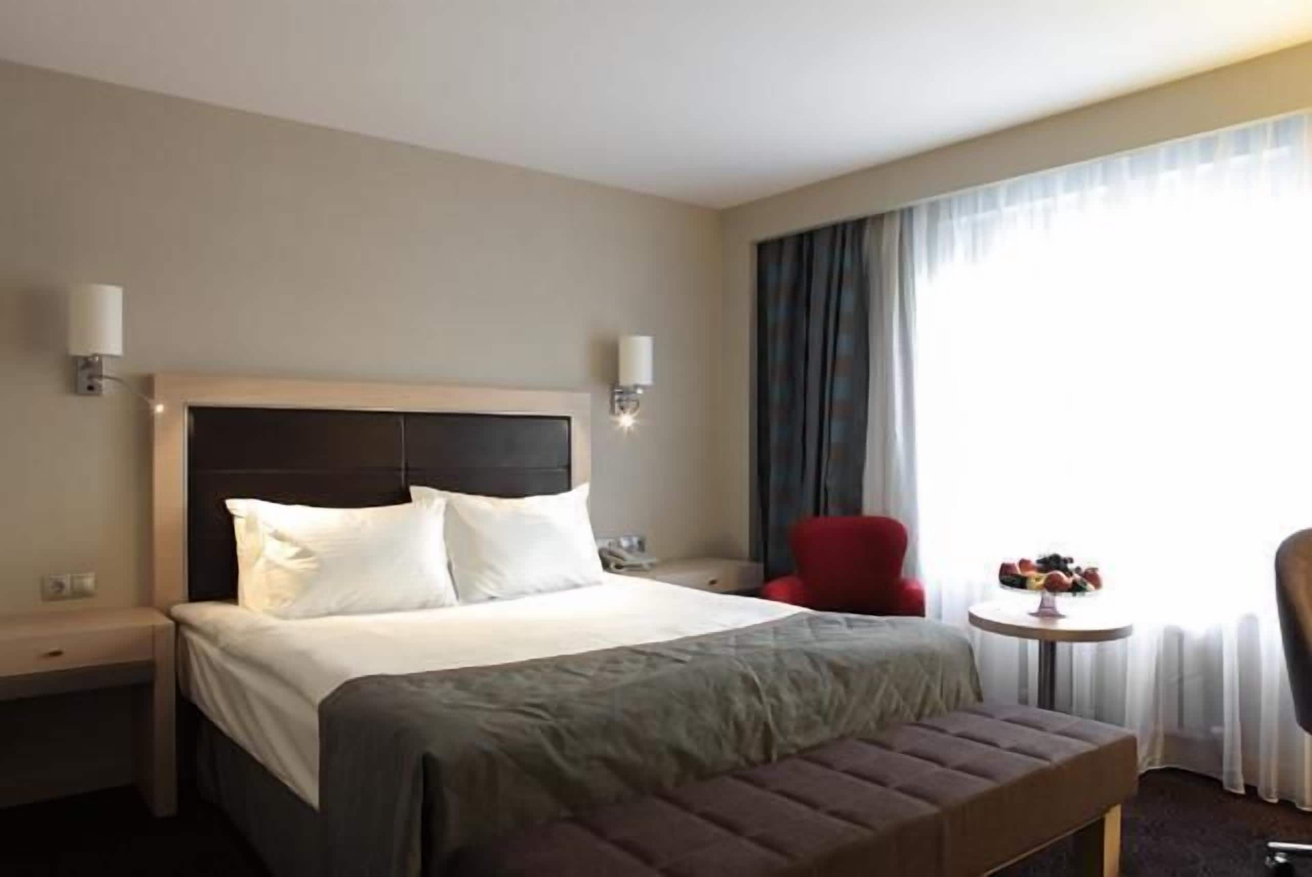 Habitación de hotel con cama, una silla roja, una mesita con fruta y una ventana con cortinas.
