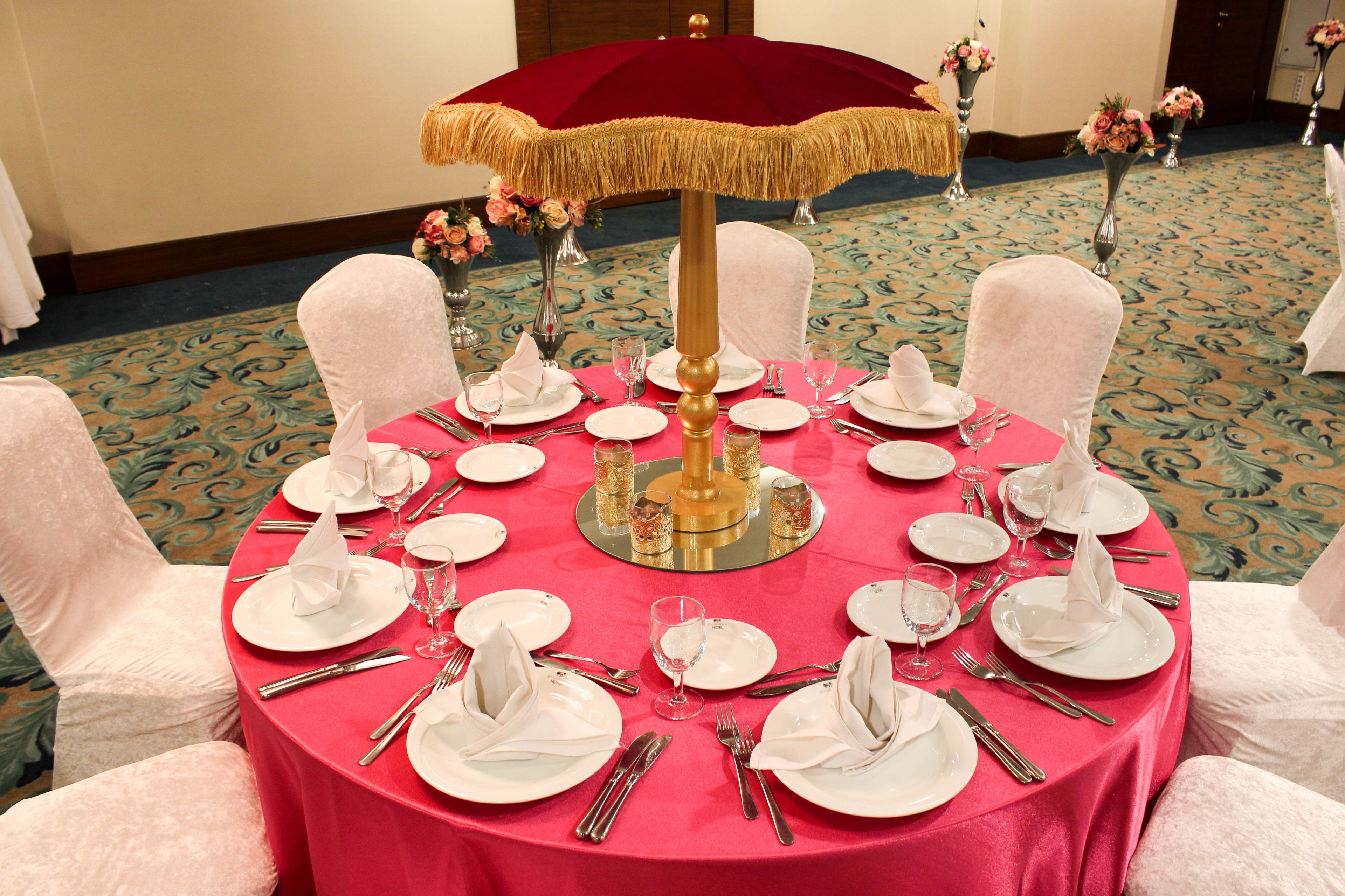 A round table set for a formal event with white chairs, a red tablecloth, and a gold umbrella centerpiece.