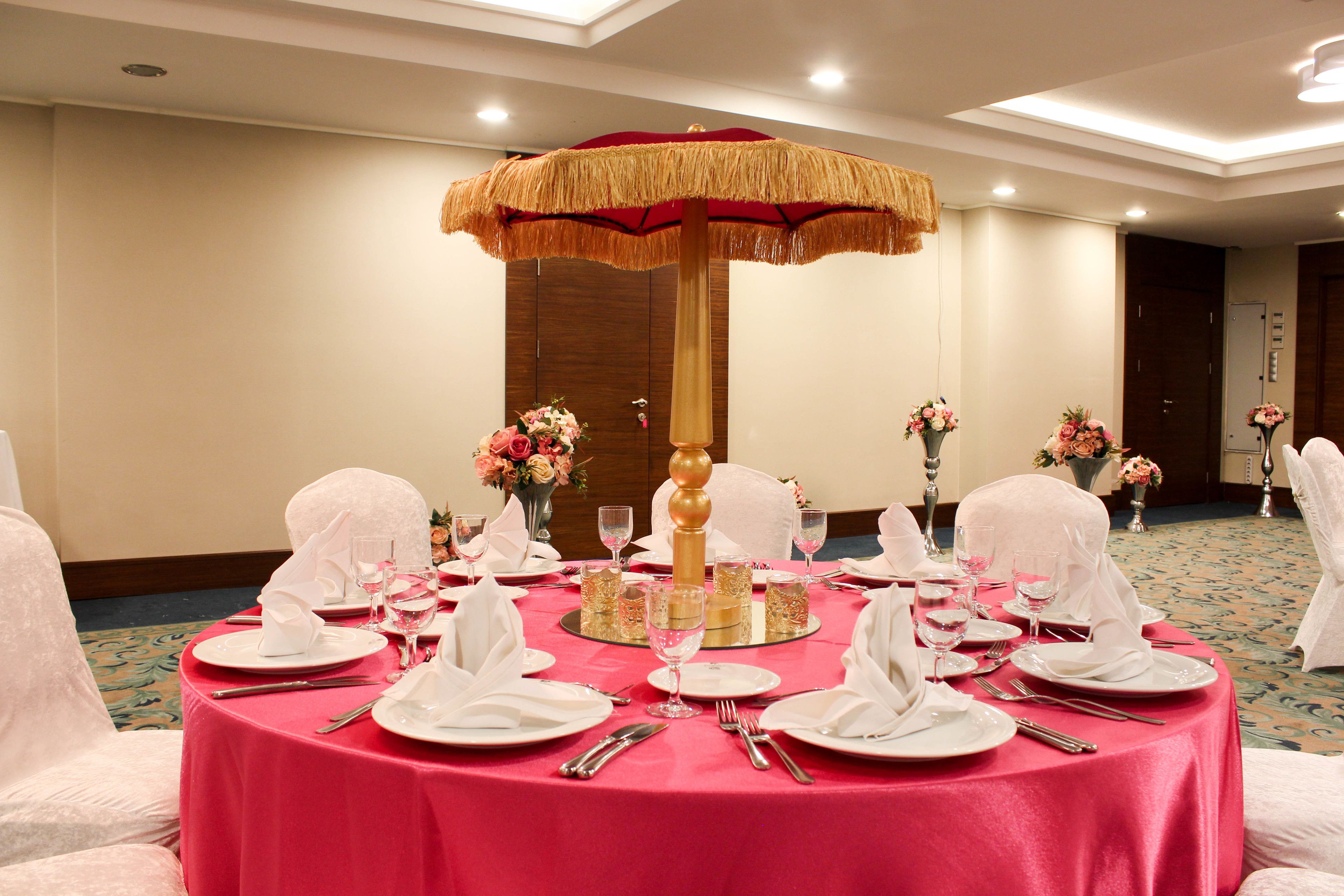 Una mesa redonda dispuesta para un evento formal, con mantel rojo, vajilla blanca y un centro de mesa decorativo con un paraguas.
