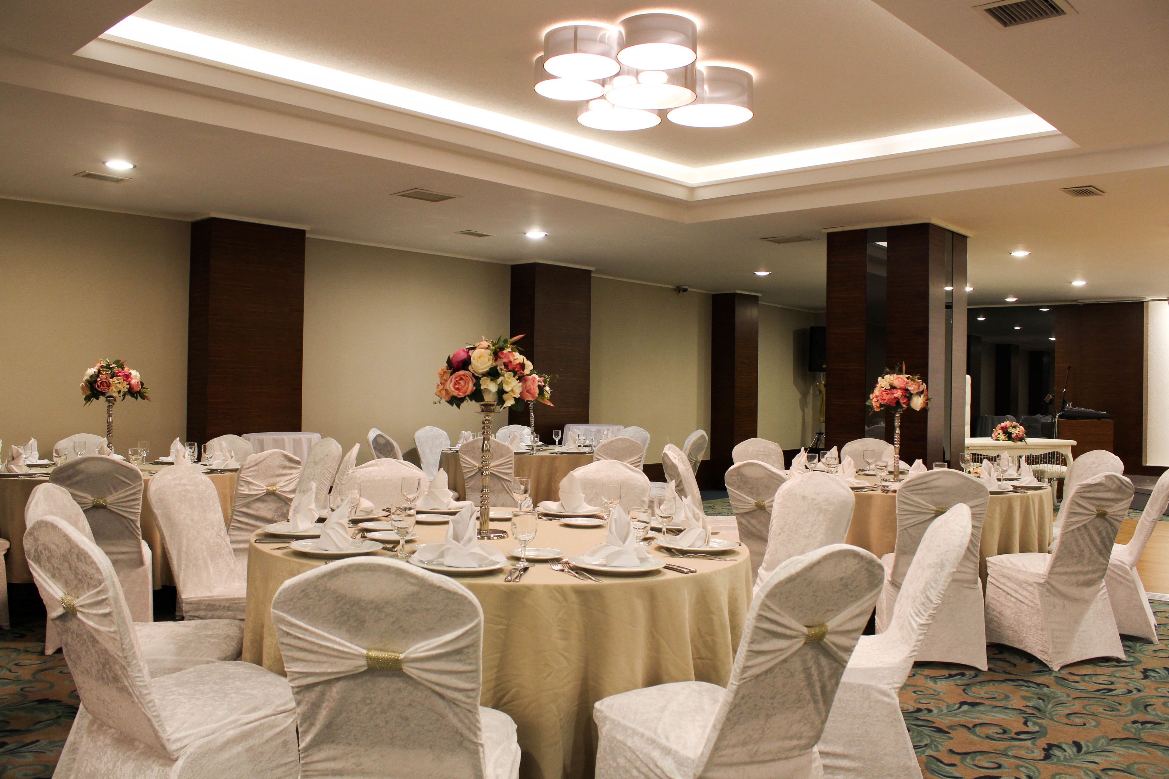 Un salón de banquetes con mesas redondas, sillas blancas y centros de mesa con flores.
