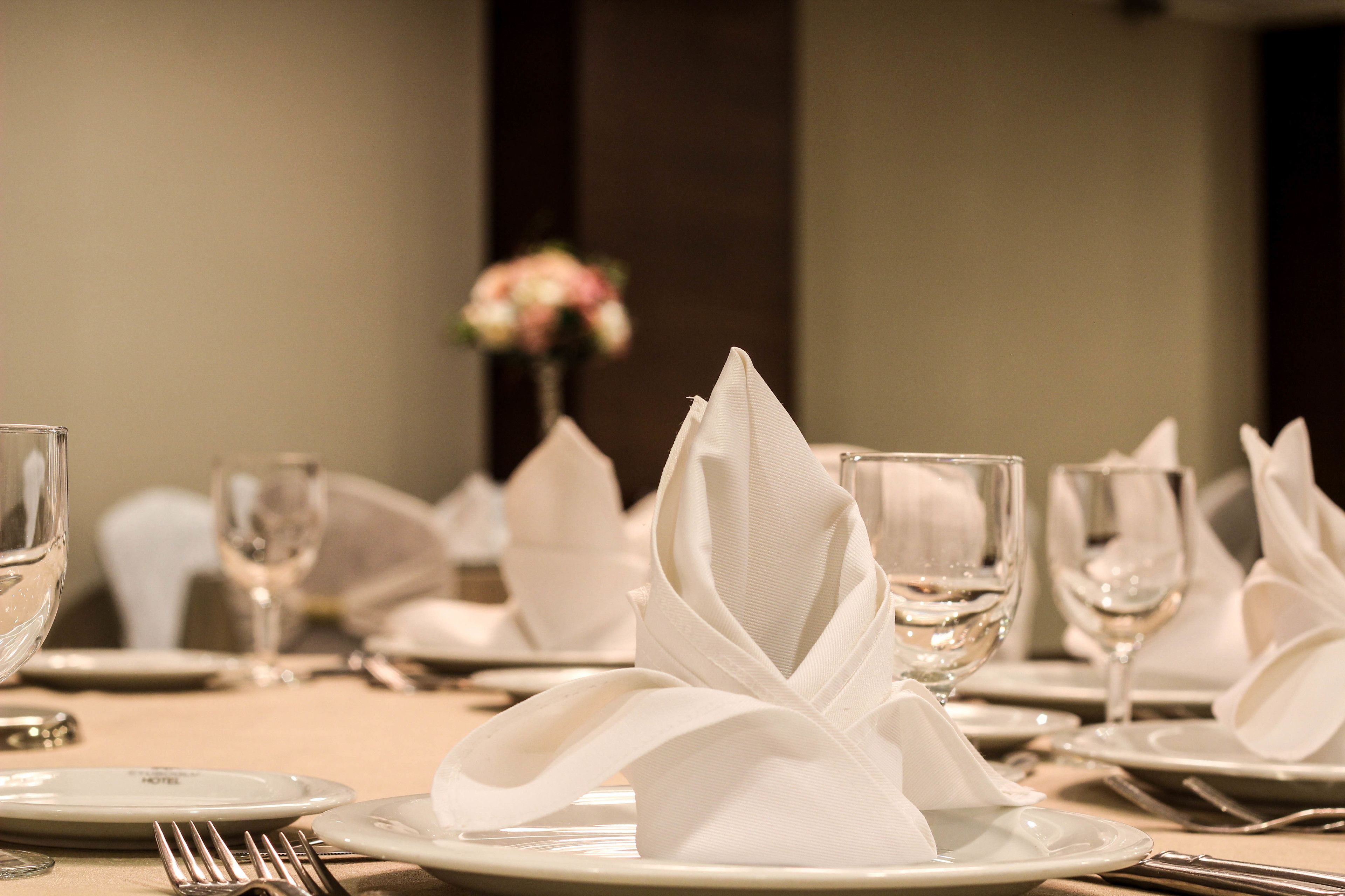 Une table dressée pour le dîner, avec des serviettes blanches pliées en forme de cygnes, des verres transparents et un vase rempli de fleurs en arrière-plan.