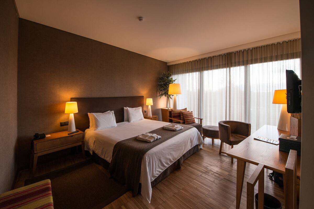 A hotel room with a large bed, two bedside lamps, a desk with a chair, and a window with curtains.