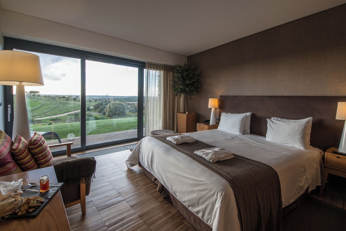 A hotel room with a large bed, a desk with a chair, a view of the countryside, and a large window with curtains.