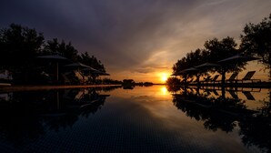 Un atardecer tranquilo sobre un estanque que refleja el cielo, con sillones de descanso y sombrillas, todo rodeado de árboles.