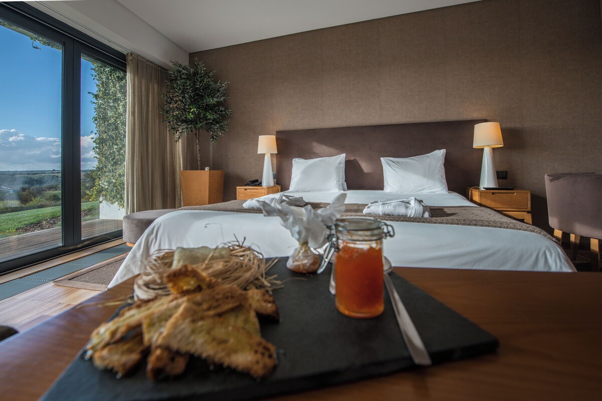 A hotel room with a large bed, two bedside tables with lamps, a view of the outdoors, and a breakfast setup on the table.