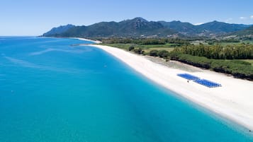 Vue aérienne d’une plage aux eaux turquoise et limpides, bordée d’un rivage de sable blanc, avec des montagnes en arrière-plan.