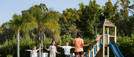 Un parque infantil con tobogán y estructuras para escalar, rodeado de árboles y césped.