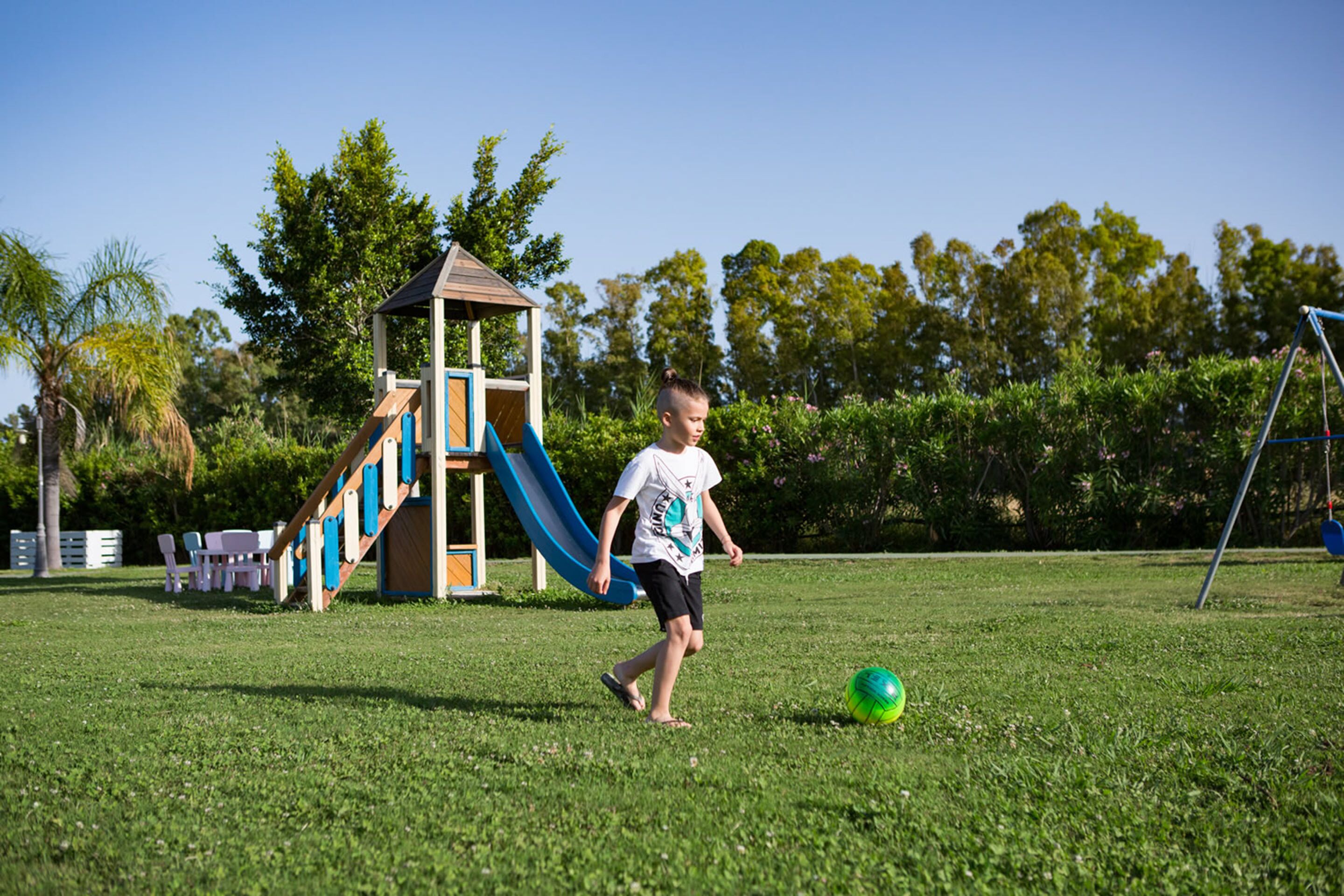 Niño jugando al fútbol en un parque con un área de juegos y árboles al fondo.