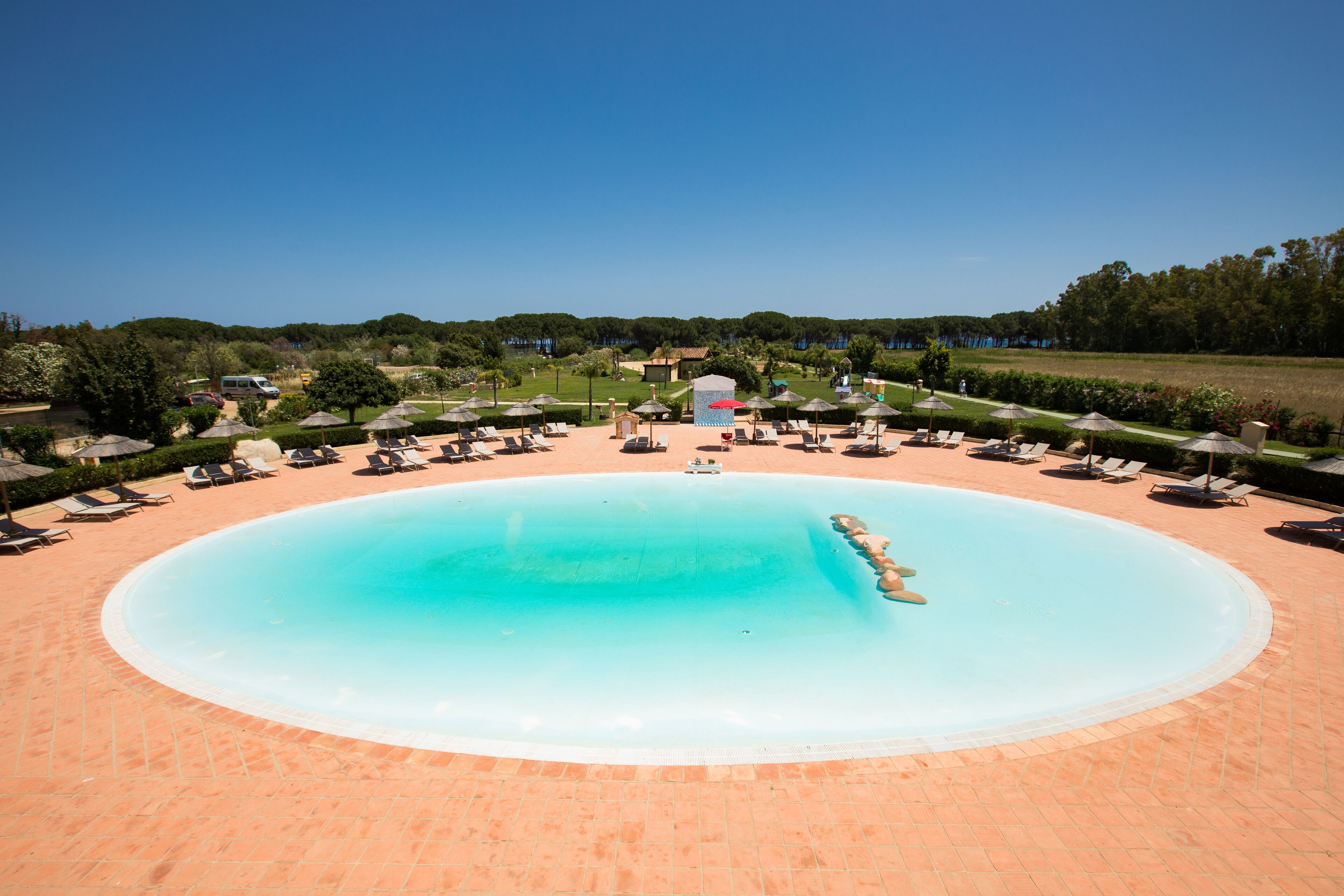 A large oval swimming pool surrounded by lounge chairs and umbrellas, with a clear sky above.
