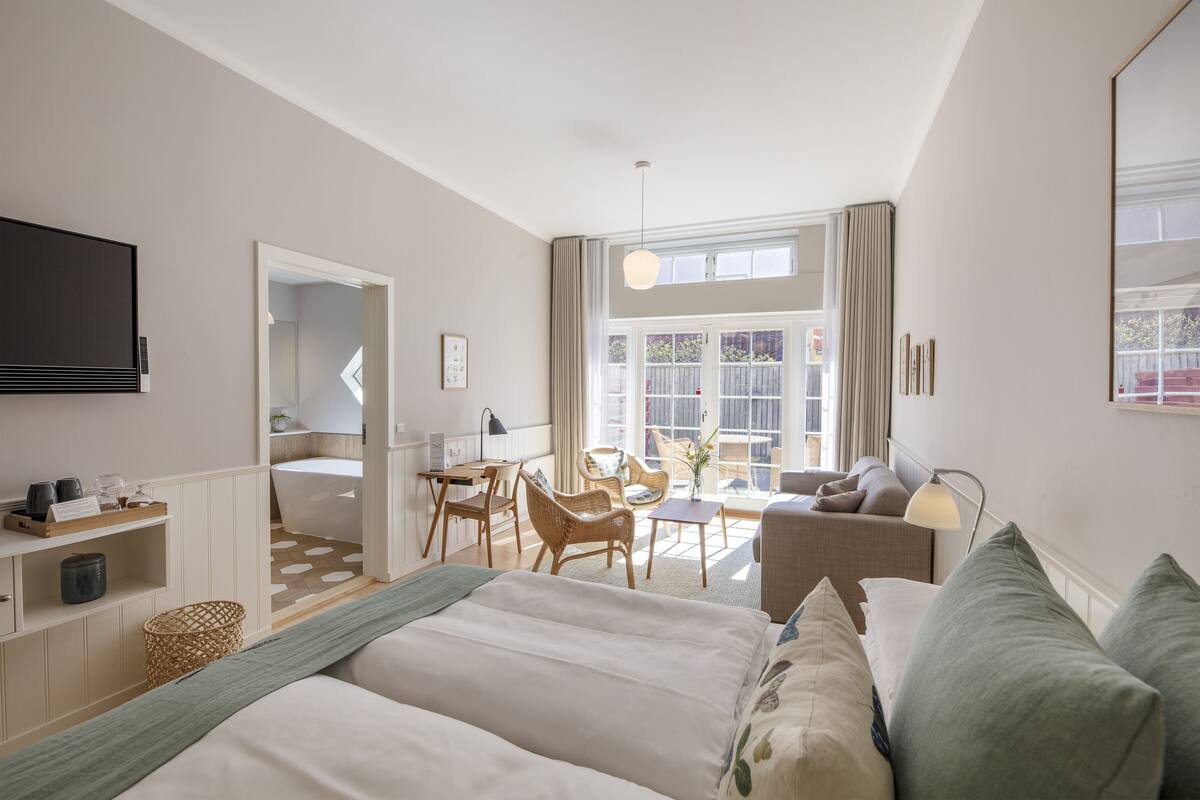 A modern hotel room with a bed, a desk, a chair, a sofa, and a TV. There is a window with curtains and a view of the outside.