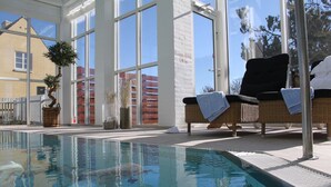 An indoor swimming pool with clear glass walls, wicker lounge chairs, and a potted plant. - Ruths Hotel (Skagen)