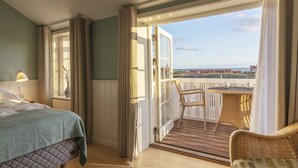 A hotel room with a bed, a window, a balcony with wicker furniture, and a view of the sea. - Ruths Hotel (Skagen)