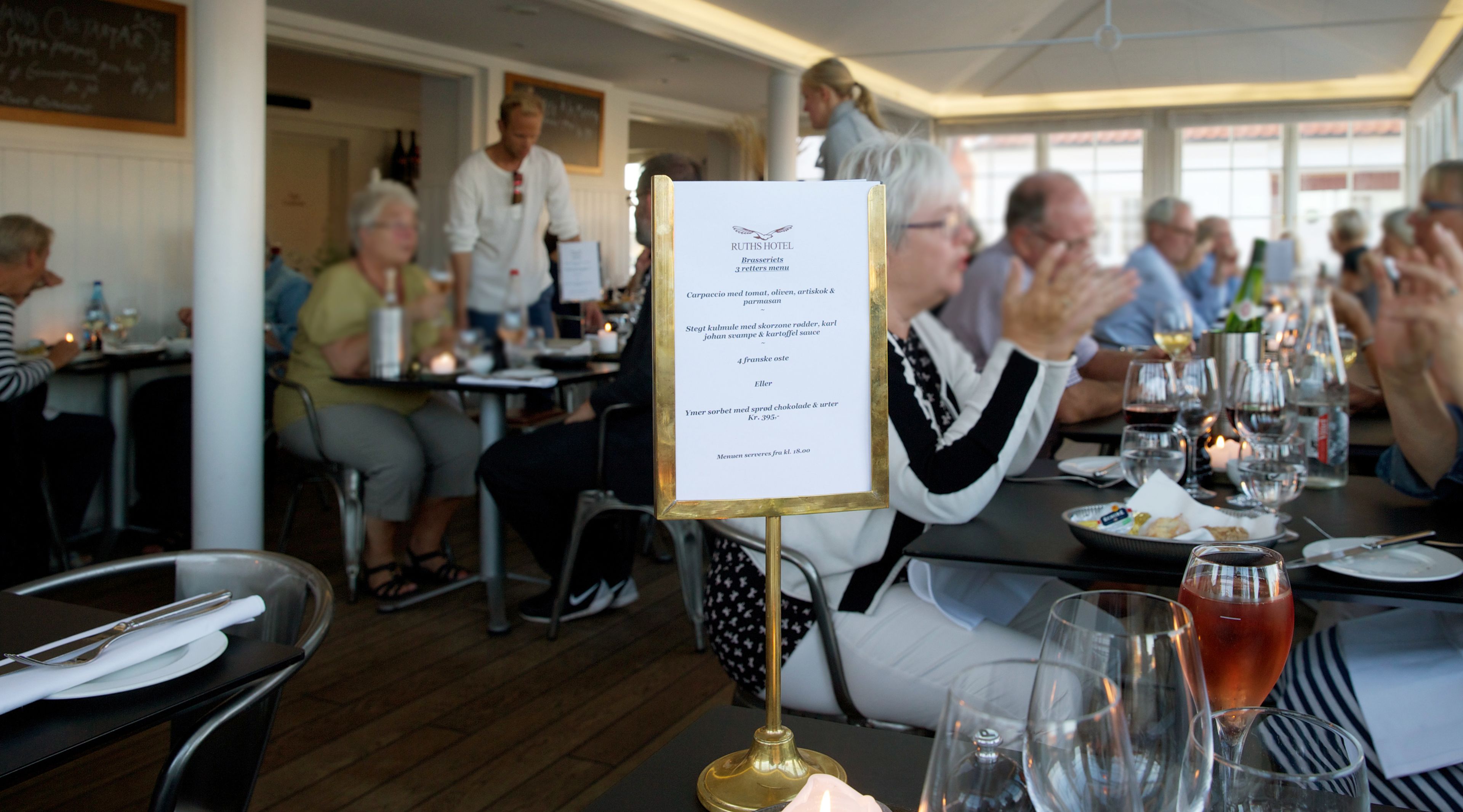 A restaurant setting with tables set for dining, a menu stand, and patrons seated at tables.