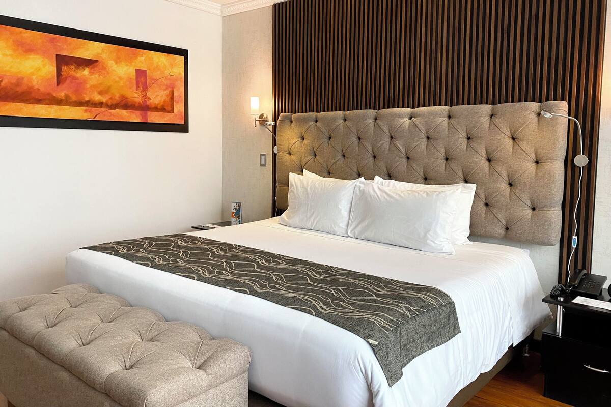 A hotel room with a large bed, a tufted headboard, a bedside lamp, a bedside phone, and a bench.
