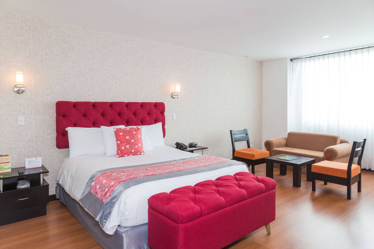 A hotel room with a large bed, a red upholstered bench, a sofa, and a chair. There is a small table and a nightstand with a phone and a lamp.
