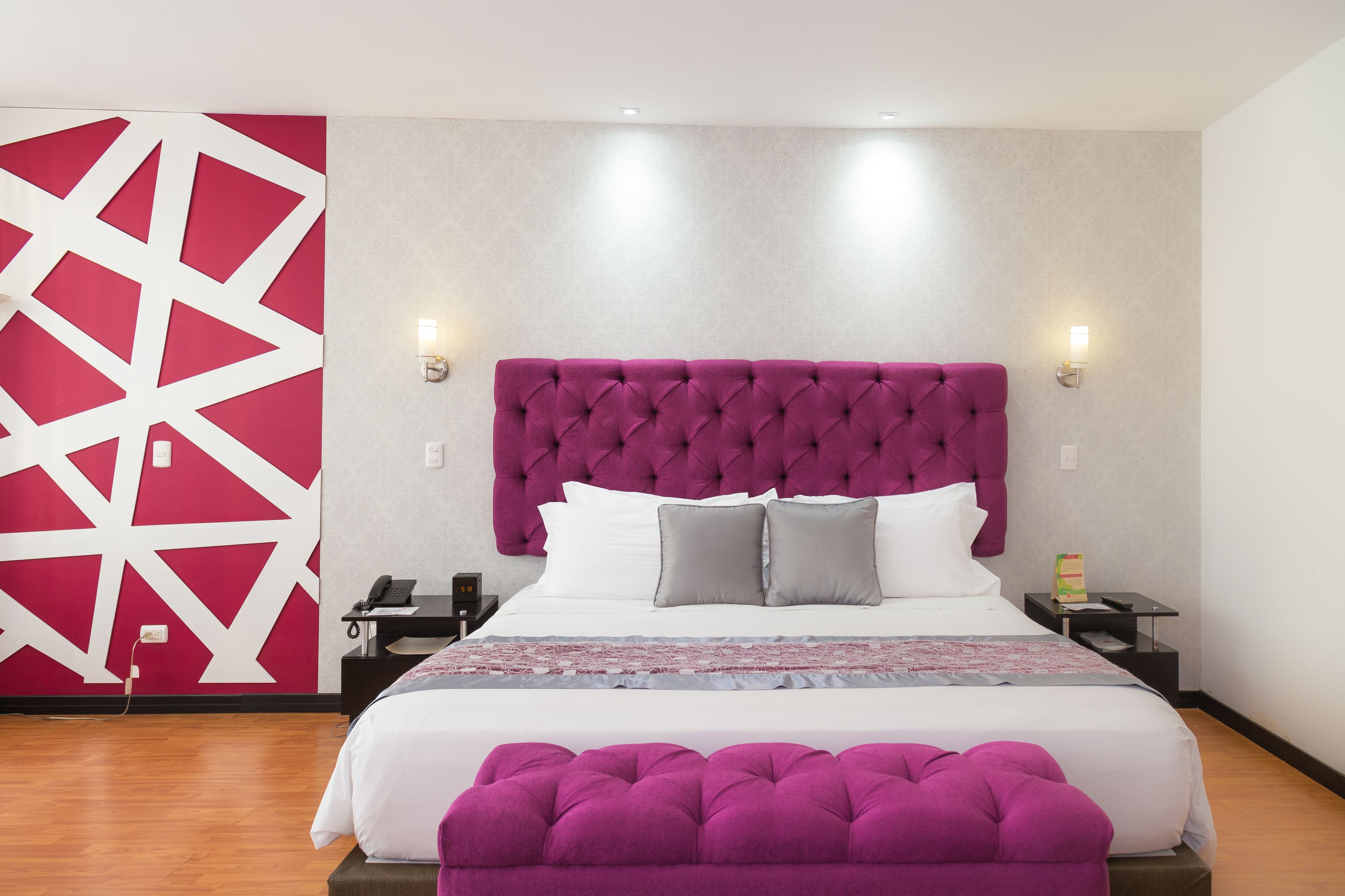 A modern hotel room with a large bed, a tufted headboard, two bedside lamps, a nightstand, and a decorative wall panel.
