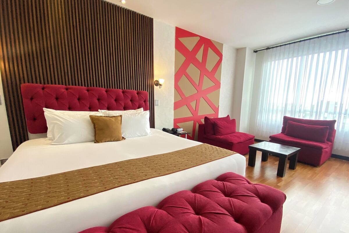A hotel room with a large bed, red upholstered chairs, a small table, and a decorative wall panel.