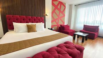 Habitación de hotel con una cama grande, sillones tapizados en rojo, una mesita pequeña y un panel decorativo en la pared.