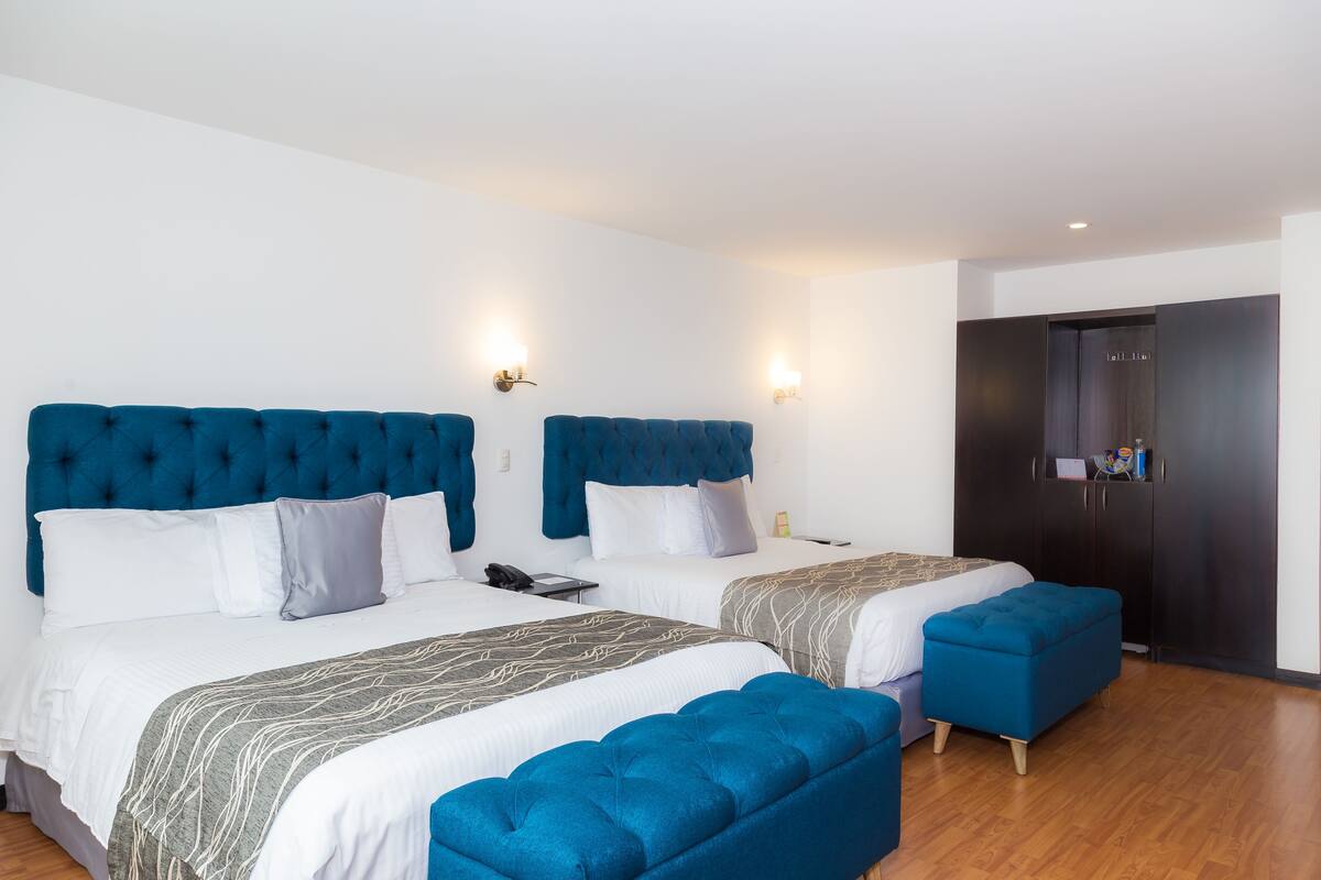 A hotel room with two beds, each with a blue headboard, white bedding, and a grey patterned throw. There is a wooden floor and a dark-colored wardrobe.