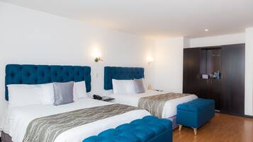 Habitación de hotel con dos camas, cada una con cabecero azul, ropa de cama blanca y una manta gris estampada. El suelo es de madera y hay un armario de color oscuro.