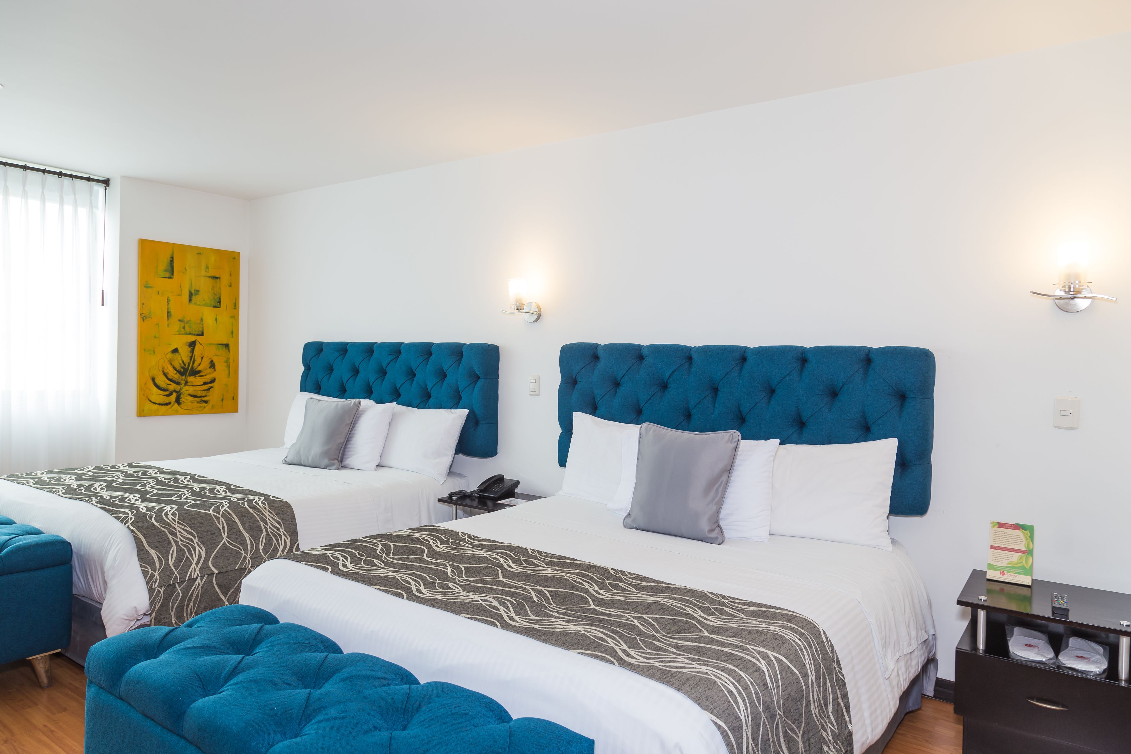 A hotel room with two beds, each with a tufted headboard, white bedding, and a grey accent pillow. There is a small bedside table with a phone and a box of tissues. A yellow abstract painting is on the wall, and there are two wall-mounted lights.