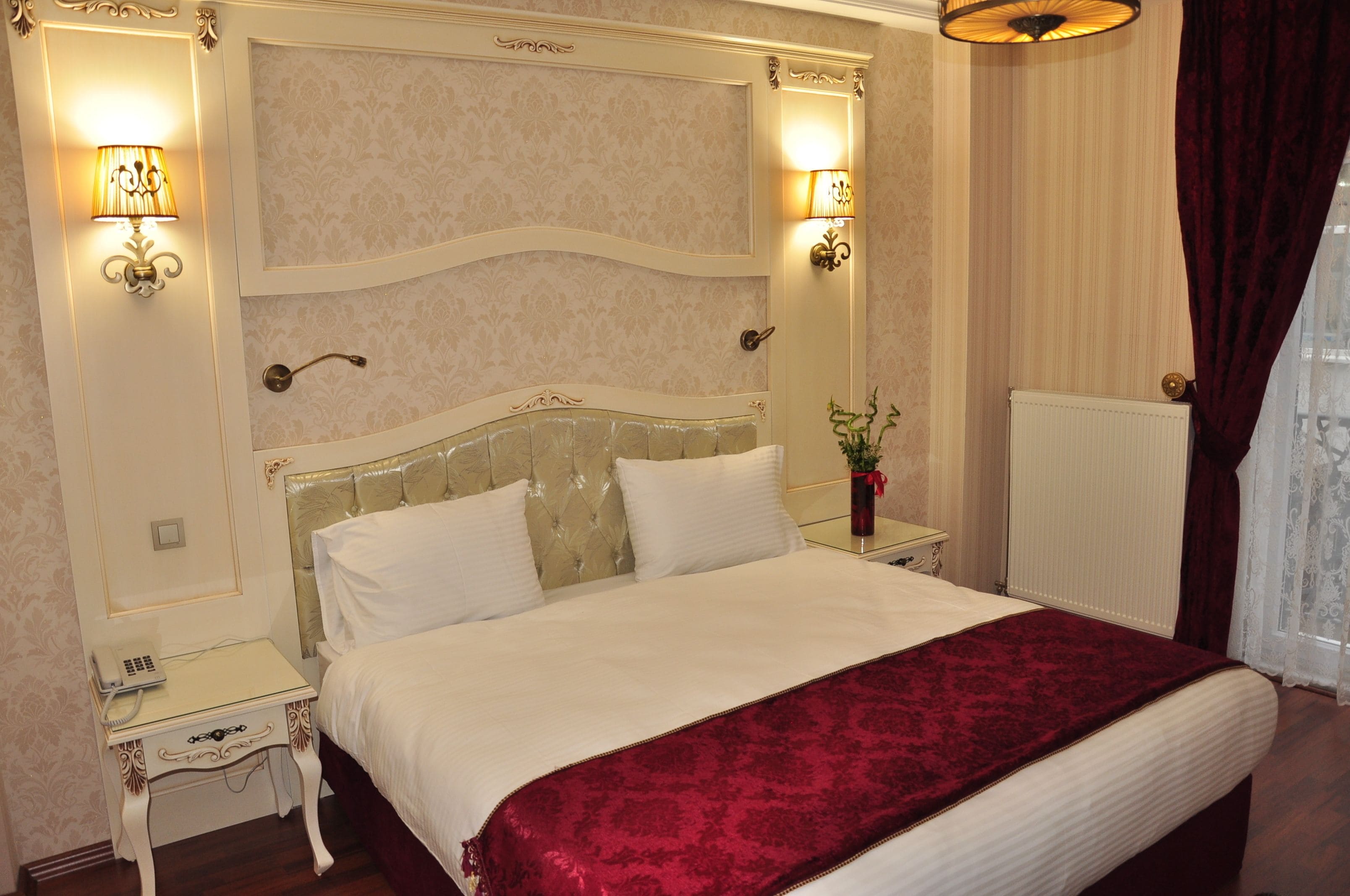 A hotel room with a large bed, two bedside tables, a nightstand, a radiator, and two wall-mounted lamps.
