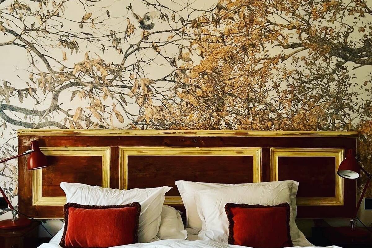 A bed with white linens and red pillows, a wooden headboard, and a wall with a tree branch and leaves mural.