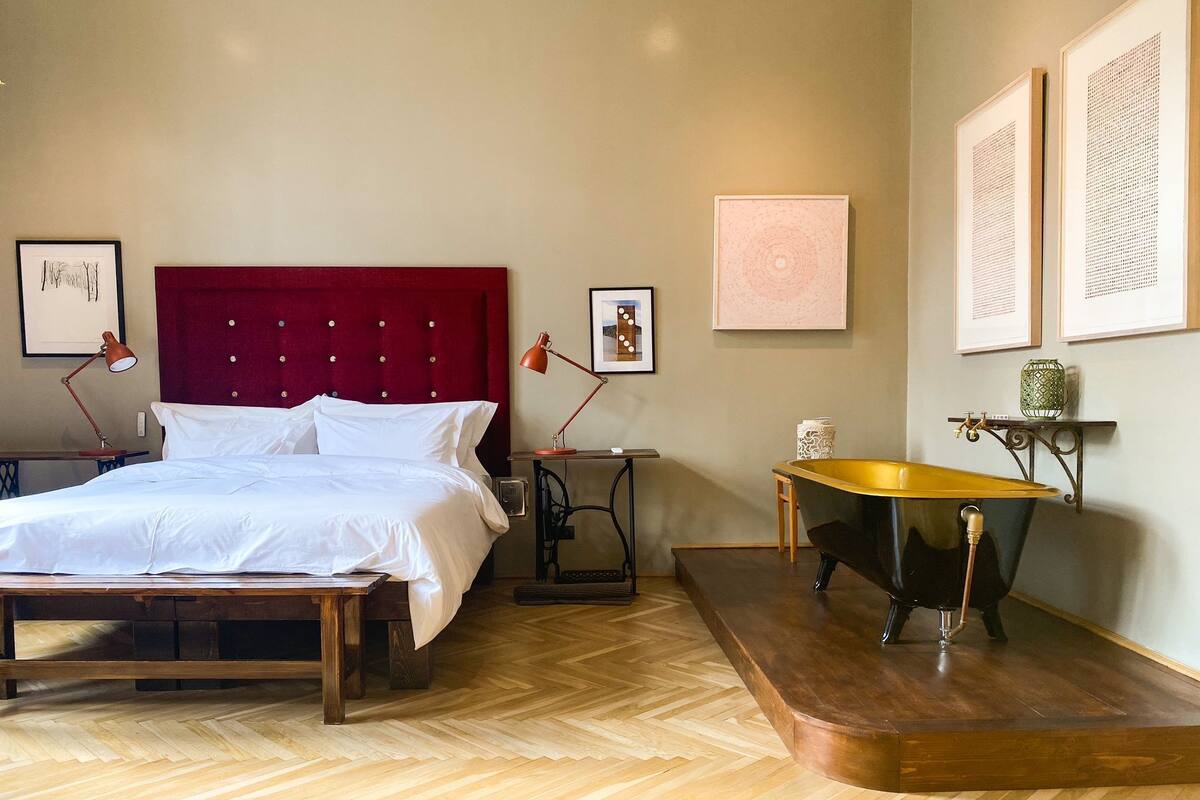 A hotel room with a red upholstered headboard, a wooden bed frame, a desk with a chair, a small sink, and a bathtub.