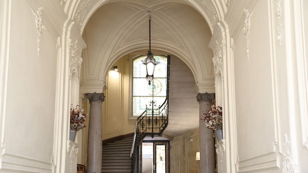 Un amplio pasillo con escaleras, columnas ornamentadas y un candelabro.
