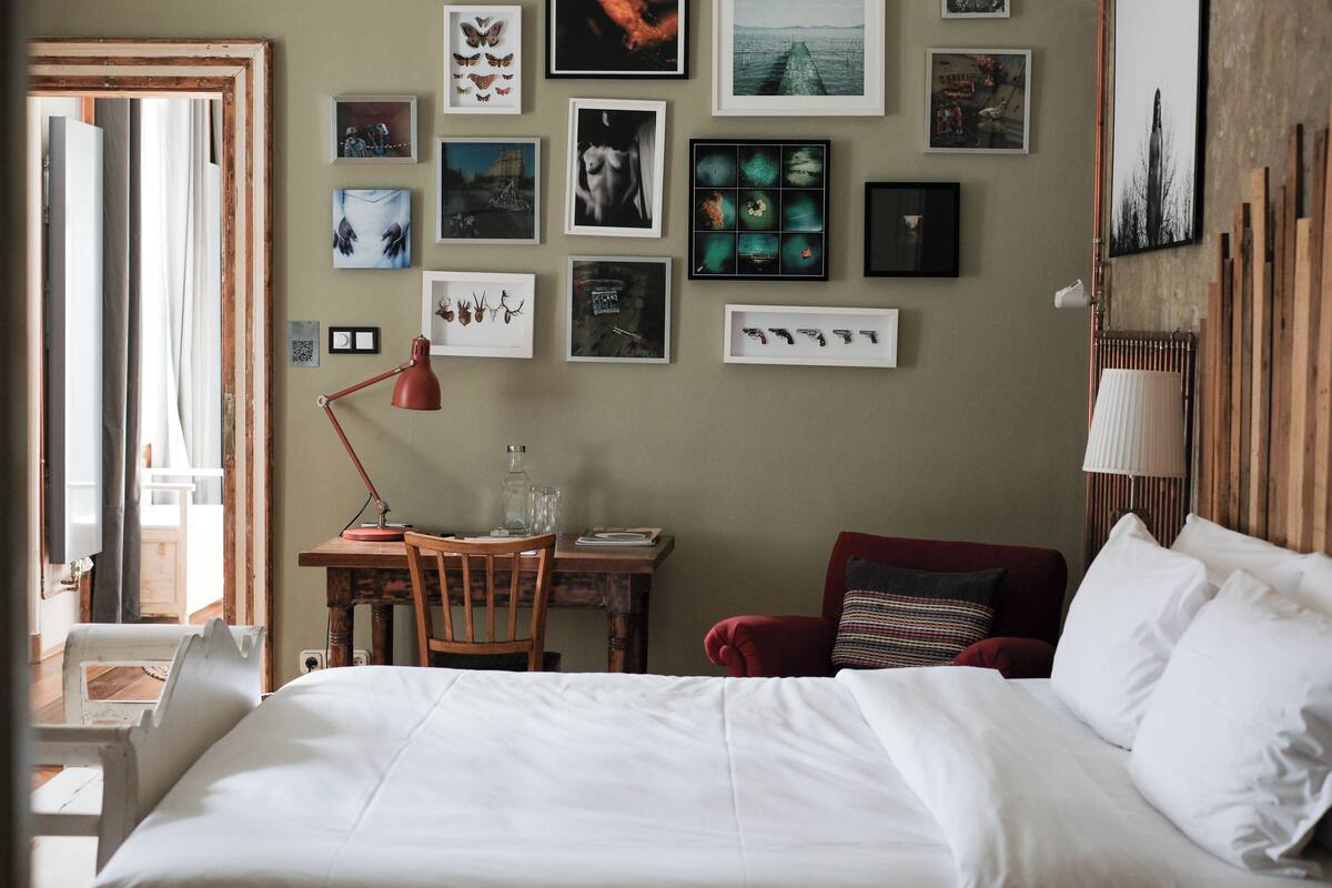 A hotel room with a bed, a desk with a chair, a red armchair, and a wall adorned with framed pictures.