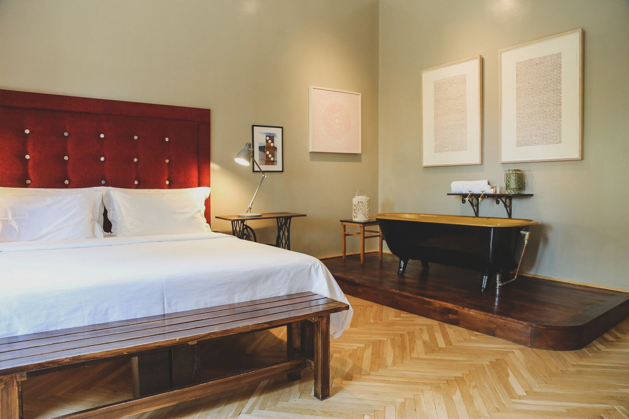 A hotel room with a large bed, a wooden bench, a black bathtub, and two framed artworks on the wall.