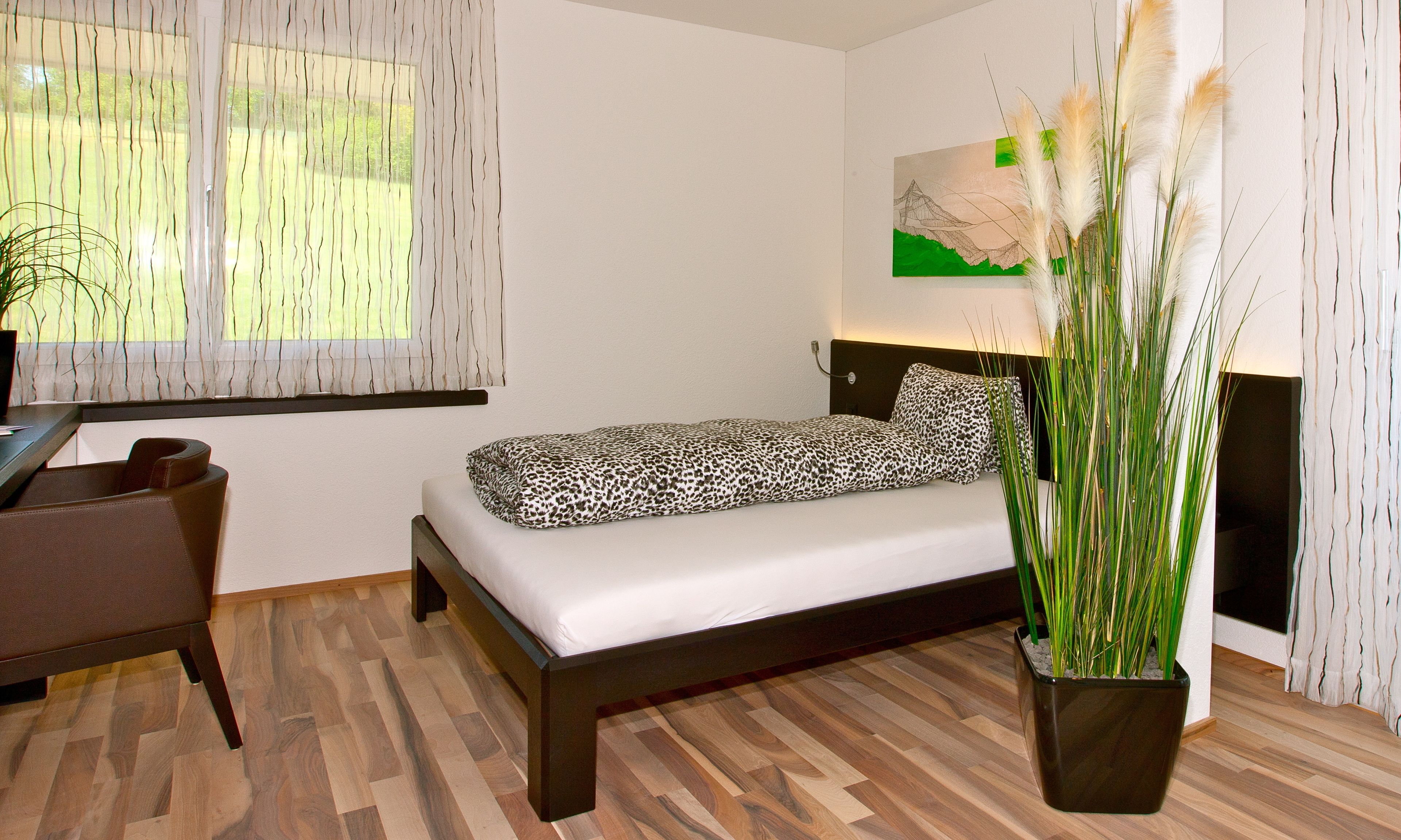 Un dormitorio moderno con suelo de madera, una cama con edredón estampado de leopardo, un escritorio con silla, un ventanal con cortinas translúcidas y una planta en maceta.