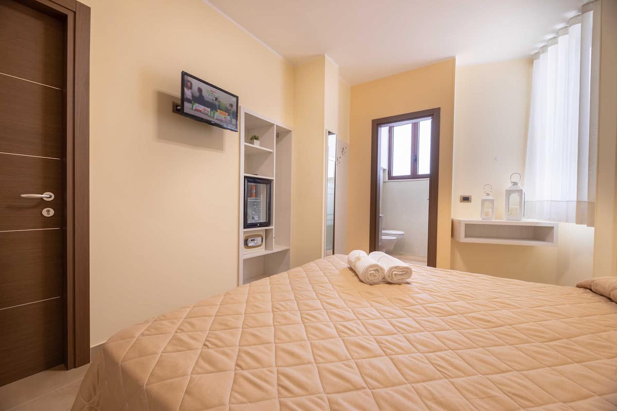 Un dormitorio con cama, televisor en un estante, puerta de baño y ventana con cortinas.